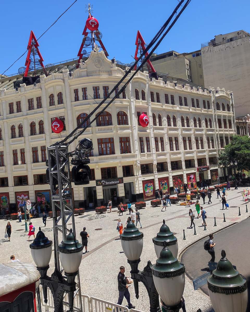 Pedestres atravessando a Rua XV de Novembro diante do Palácio Avenida, no Centro de Curitiba, num dia de céu azul, com postes antigos e prédios altos em volta.
