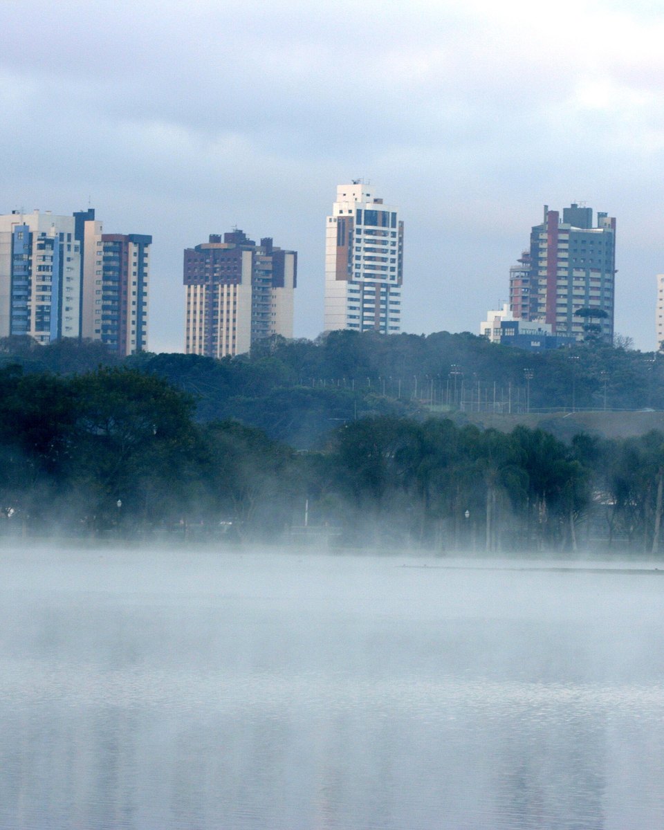 Névoa sobre o lago do Parque Barigui numa manhã fria, com prédios de Curitiba ao fundo.
