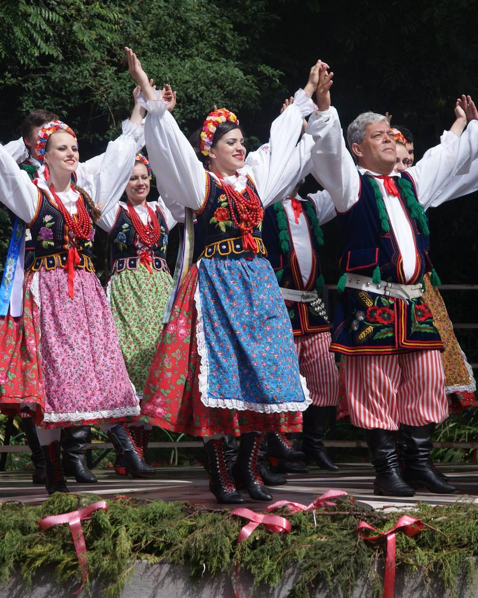 Grupo de dança folclórica polonesa com trajes típicos coloridos, de mãos dadas e braços erguidos, se apresentando ao ar livre no Bosque do Papa, em Curitiba.