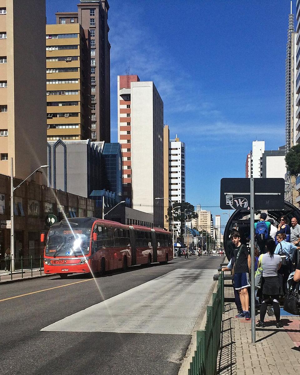 Ônibus biarticulado vermelho chegando pela canaleta exclusiva em Curitiba enquanto passageiros fazem fila na entrada da estação-tubo, entre prédios altos e céu azul.