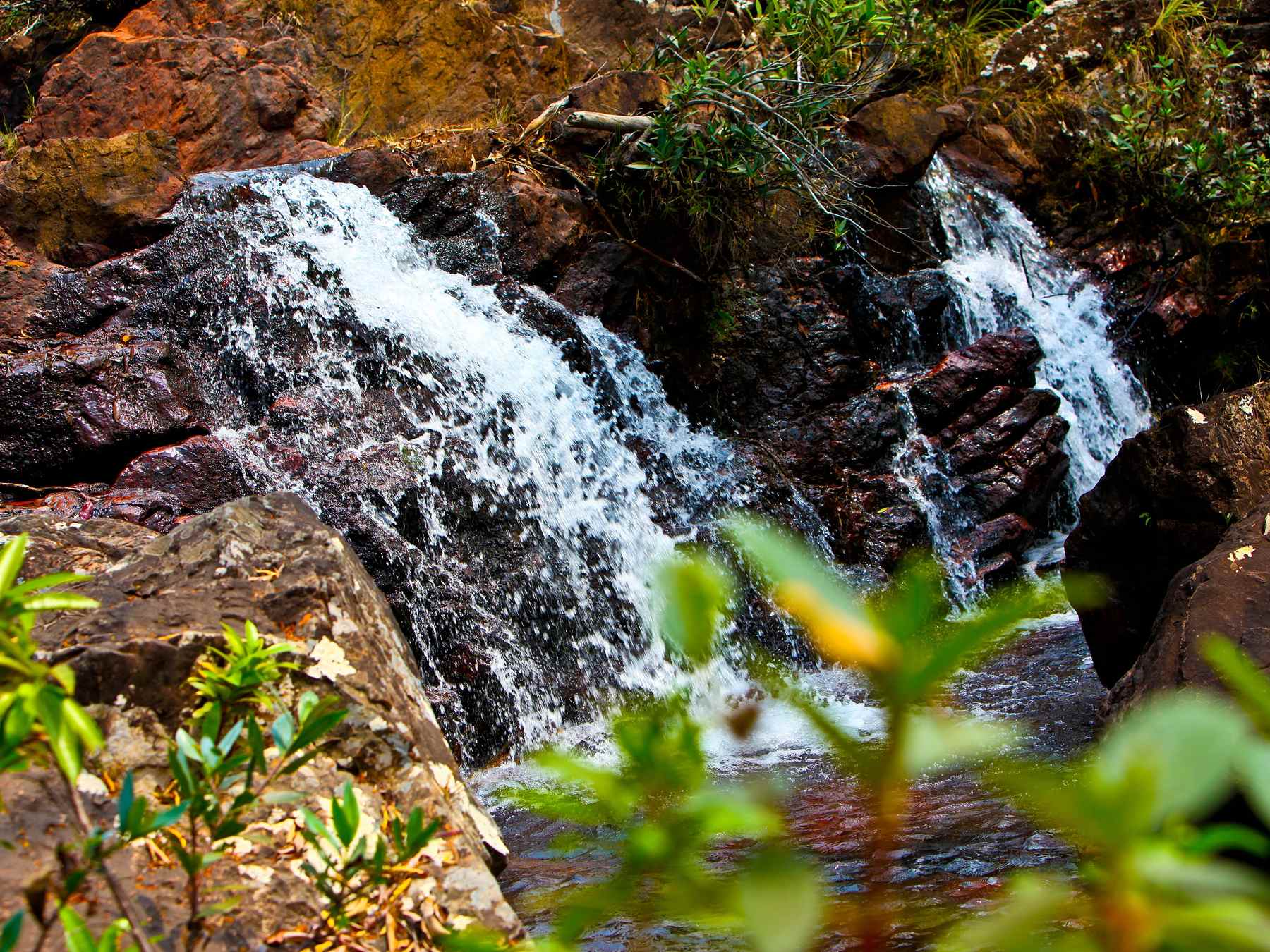 Cachoeira descendo entre paredões de pedra avermelhada da Chapada Imperial, com mata do cerrado verde em volta.