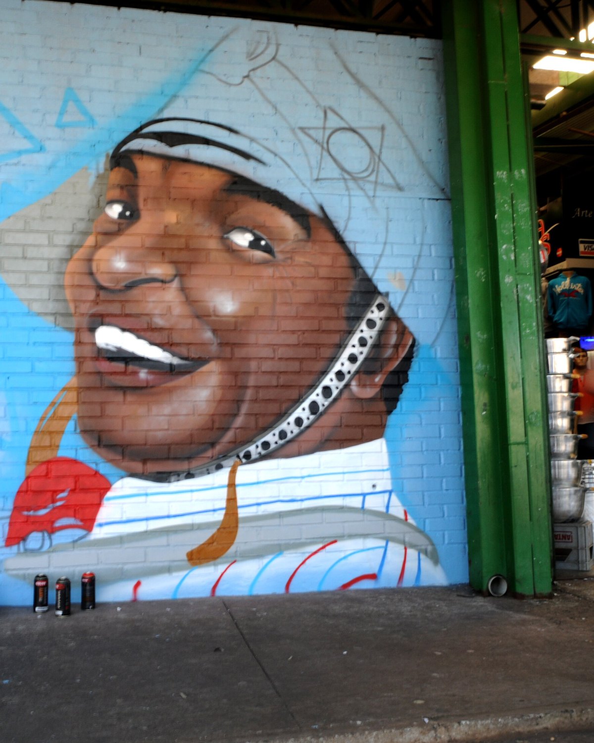 Grafite azul com o rosto sorridente de um homem de bandana pintado na parede de uma loja da Feira Central de Ceilândia, com uma cliente passando na calçada.