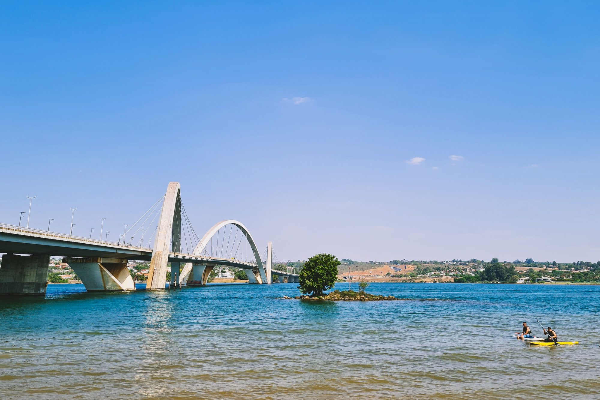 Duas pessoas num caiaque amarelo e outra em pé numa prancha de stand up nas águas do Lago Paranoá, com a Ponte JK e seus arcos brancos ao fundo, num dia de céu azul.