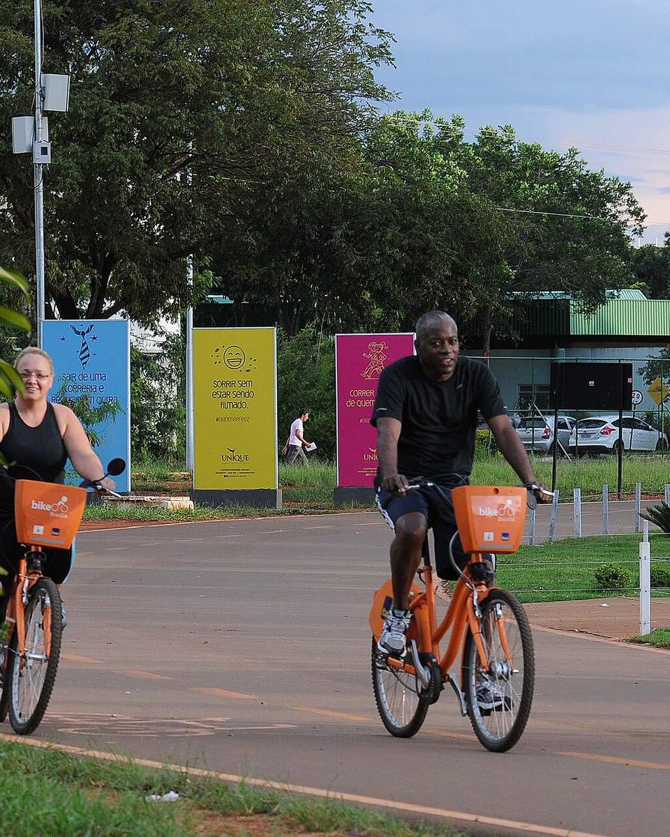 Três pessoas pedalando bicicletas laranja de aluguel numa via do Parque da Cidade, em Brasília, com árvores e painéis coloridos ao lado.
