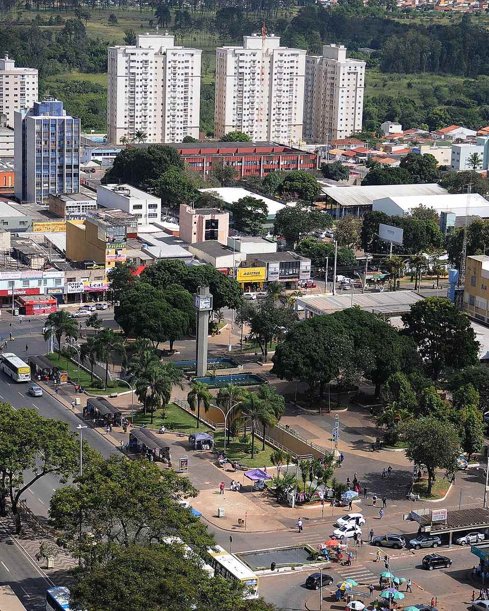 Vista do alto de Taguatinga, com uma avenida larga cheia de carros, canteiros arborizados, lojas e prédios residenciais ao fundo.
