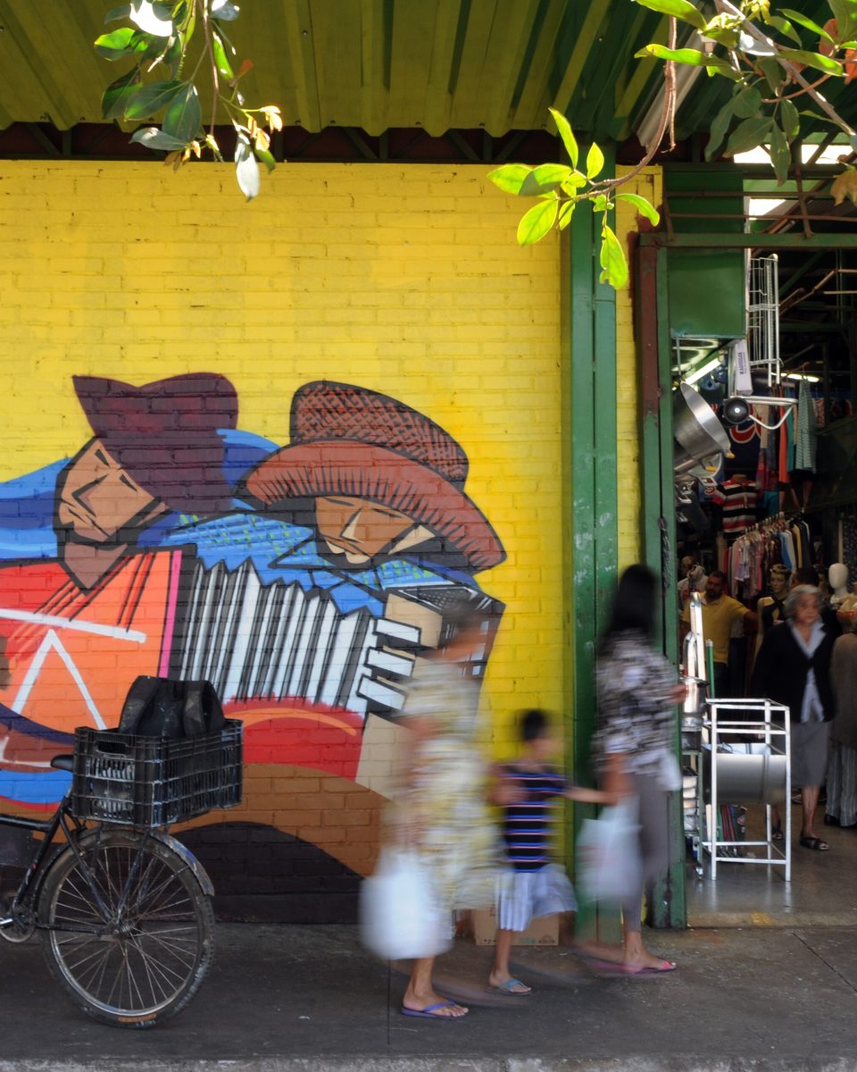 Mural colorido com figuras de sanfoneiro e vendedores nordestinos pintado na parede amarela da Feira Central de Ceilândia, com bicicletas estacionadas e clientes entrando nas lojas.