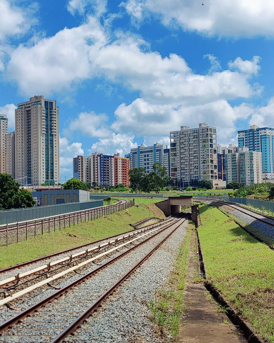 Trilhos do metrô cortando Águas Claras entre gramados, com dezenas de torres residenciais altas dos dois lados sob céu azul com nuvens.