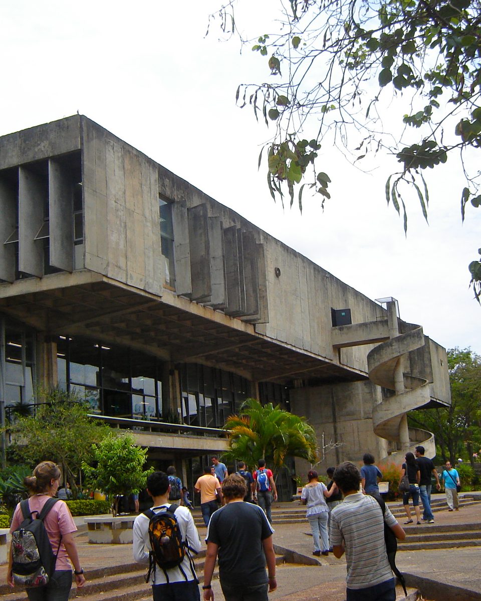 Estudantes com mochilas caminhando diante do prédio de concreto do restaurante universitário da Universidade de Brasília, com árvores em volta.