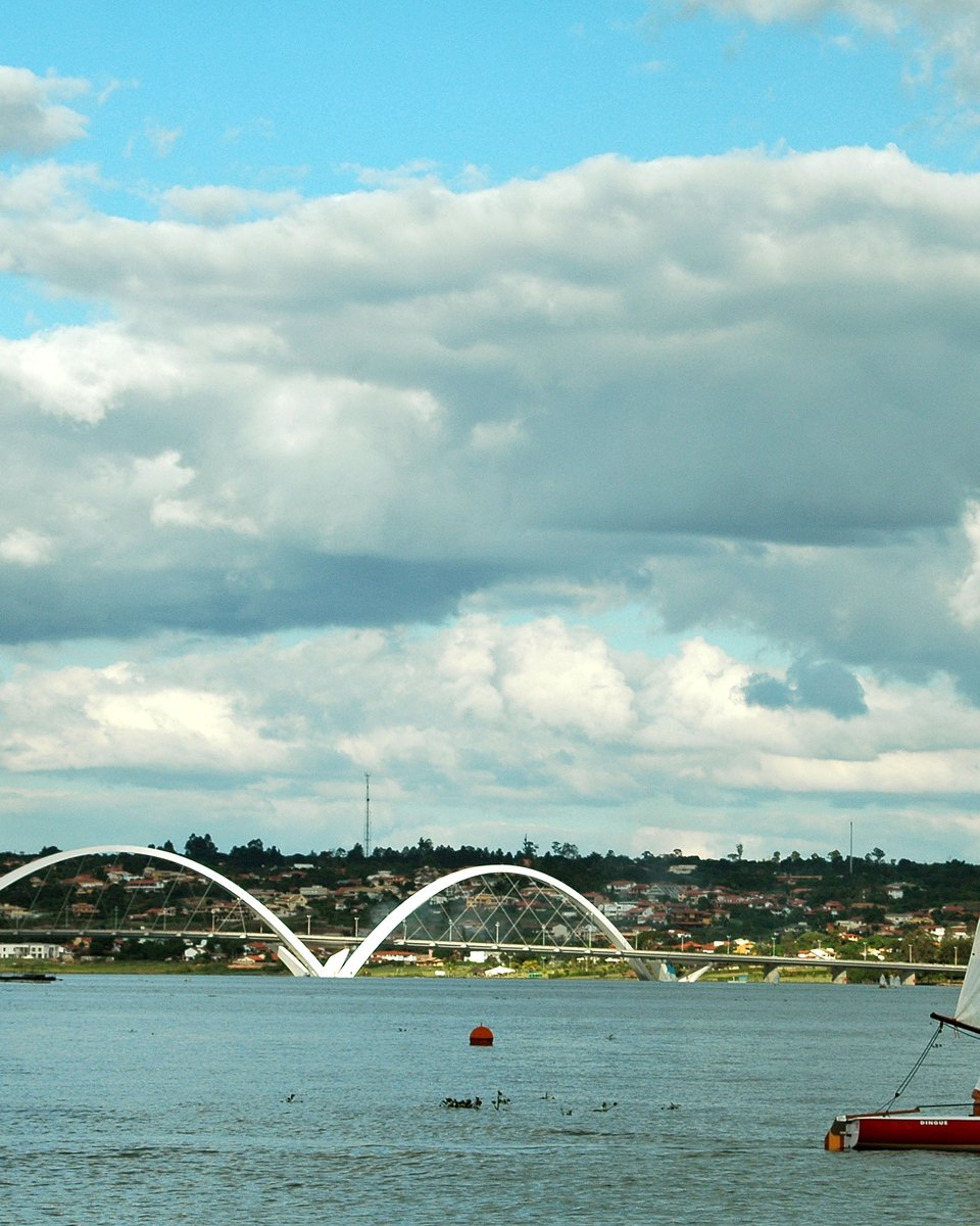 Pequeno barco a vela de pano vermelho e branco navegando no Lago Paranoá, com a Ponte JK e seus arcos ao fundo e nuvens brancas no céu.