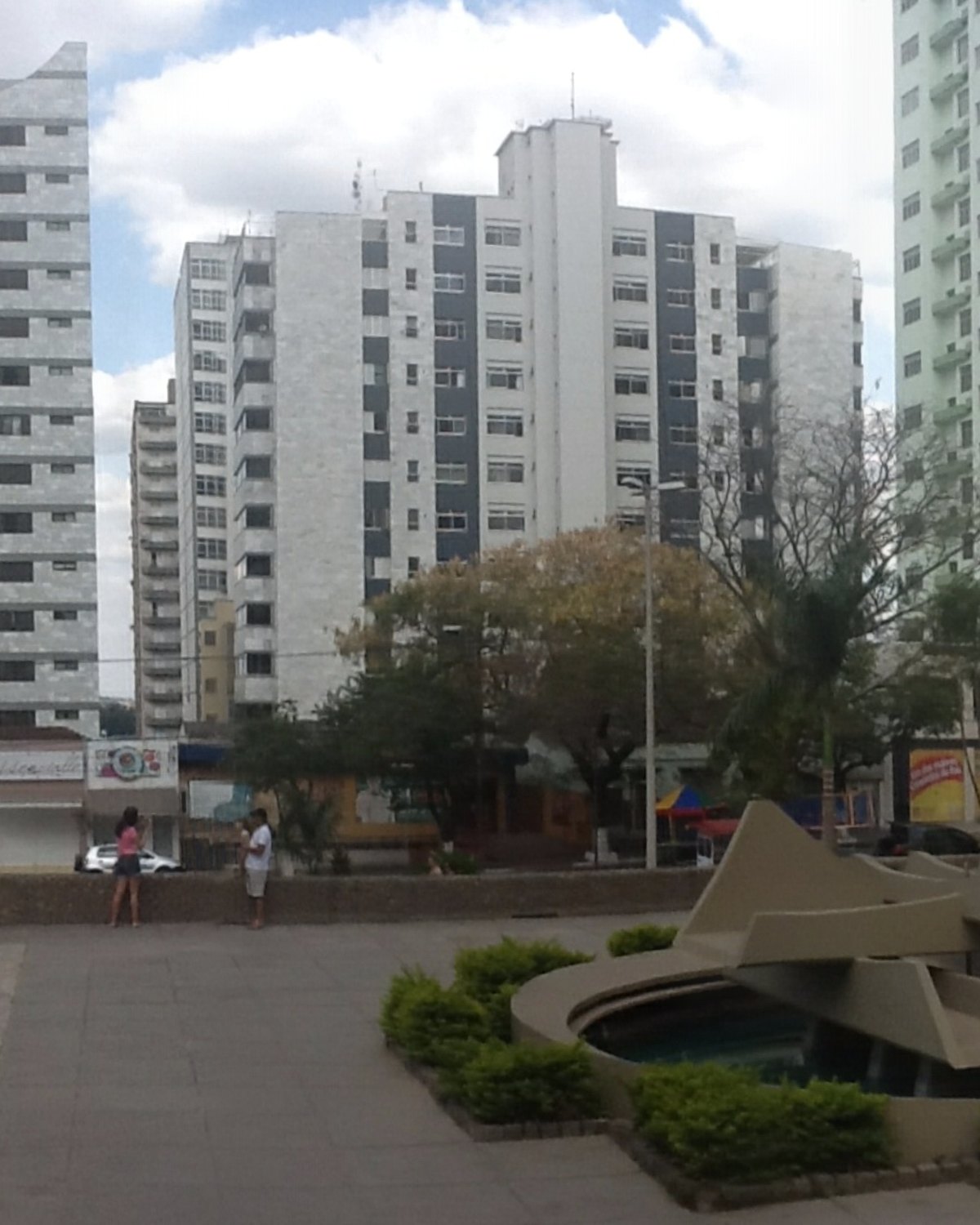Praça do Santuário, em Divinópolis, com palmeiras, canteiros e gente caminhando.
