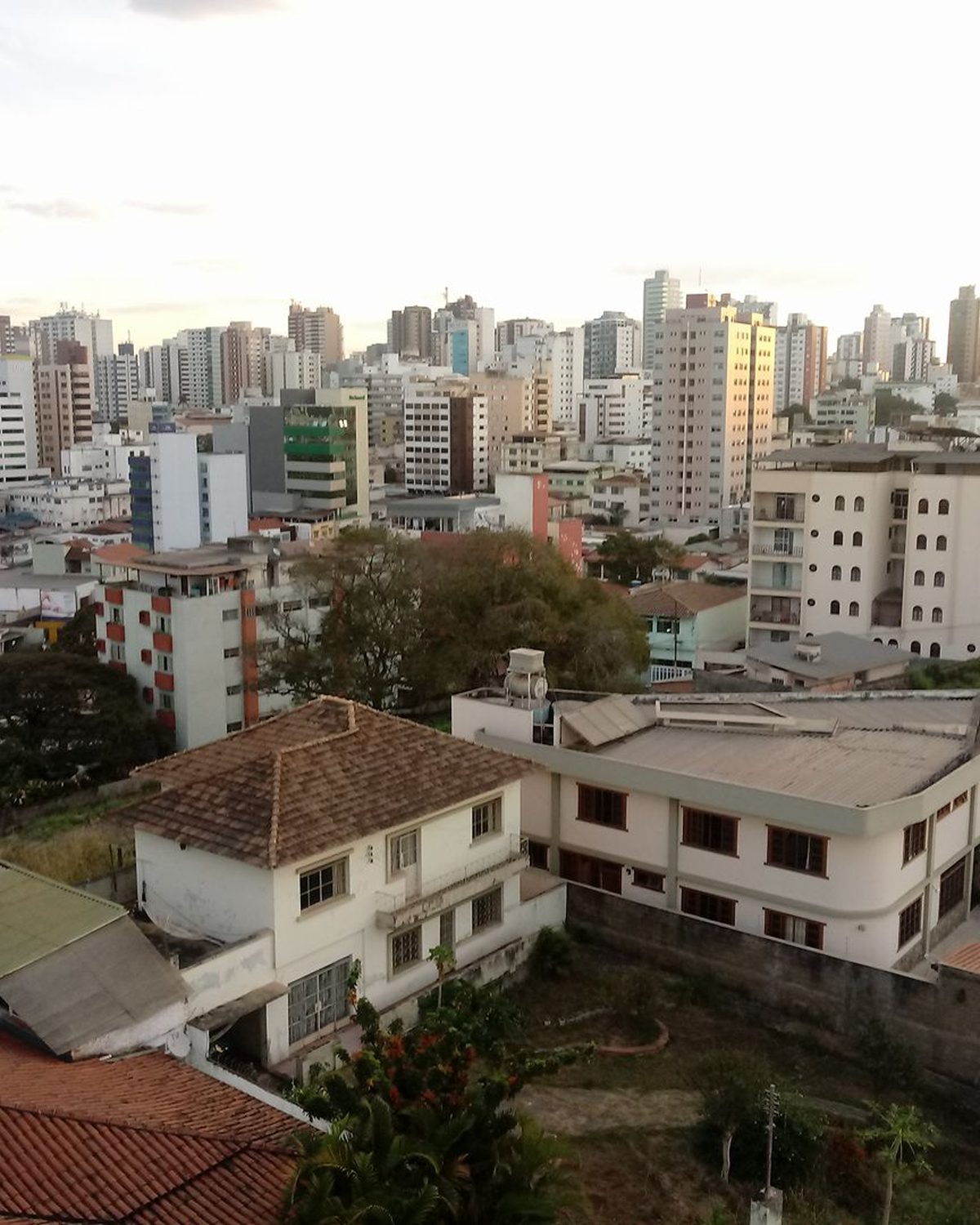 Centro de Divinópolis visto do alto, com prédios altos ao fundo, casas de telhado de cerâmica e ruas com carros estacionados em primeiro plano.