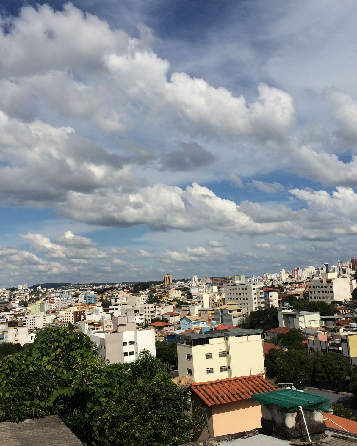 Vista ampla de Divinópolis, com casas e prédios espalhados pelos morros sob um céu azul cheio de nuvens.
