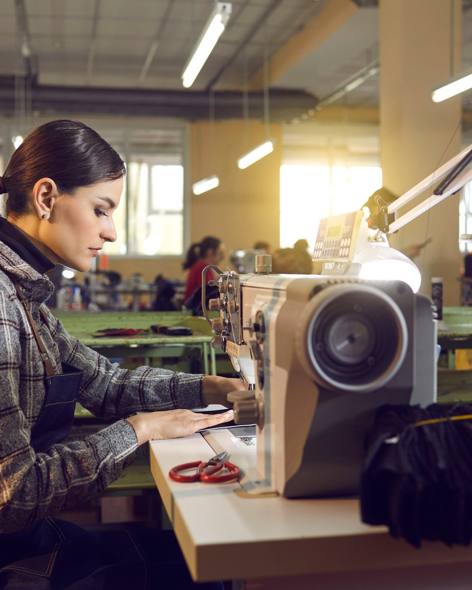 Costureira de camisa xadrez trabalhando numa máquina de costura industrial, com outras mesas de costura e colegas ao fundo, num galpão de confecção.