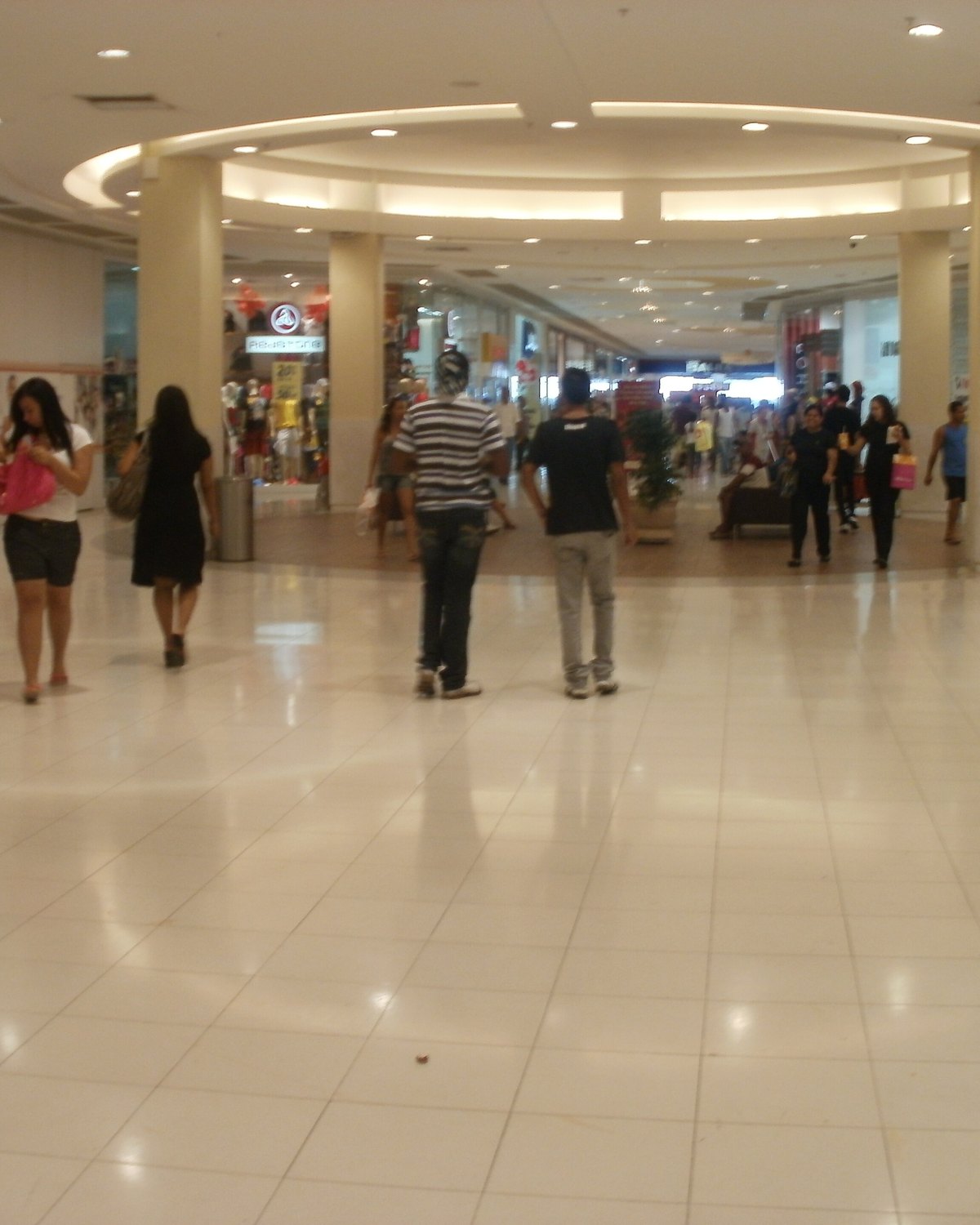 Corredor do Caxias Shopping com piso claro e pilares iluminados, com pessoas passeando entre as lojas, uma delas com sacola rosa na mão.