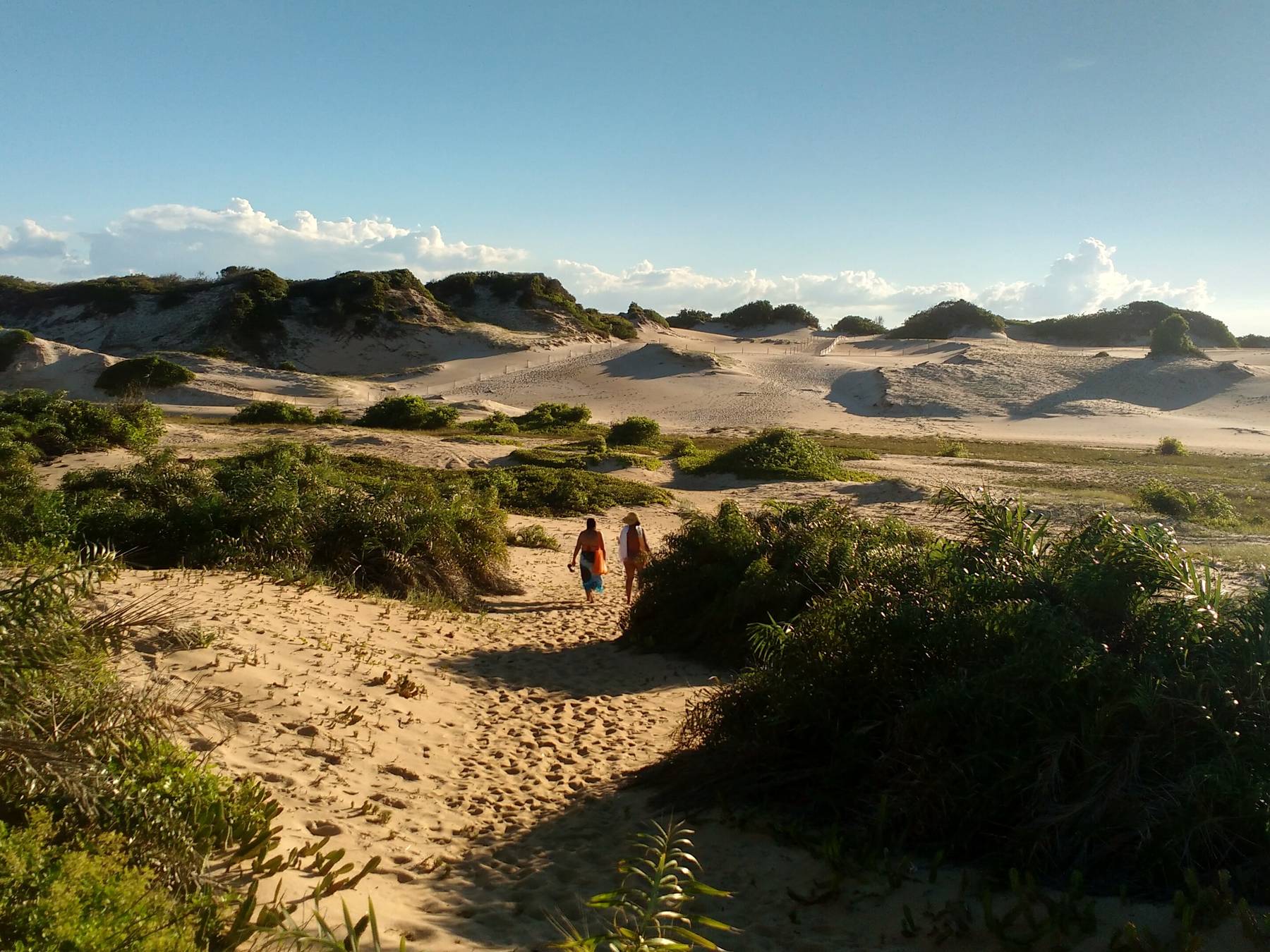 Duas pessoas subindo a trilha de areia entre a vegetação rasteira até o campo de dunas de Itaúnas, com as dunas brancas se estendendo até o horizonte sob o céu azul.