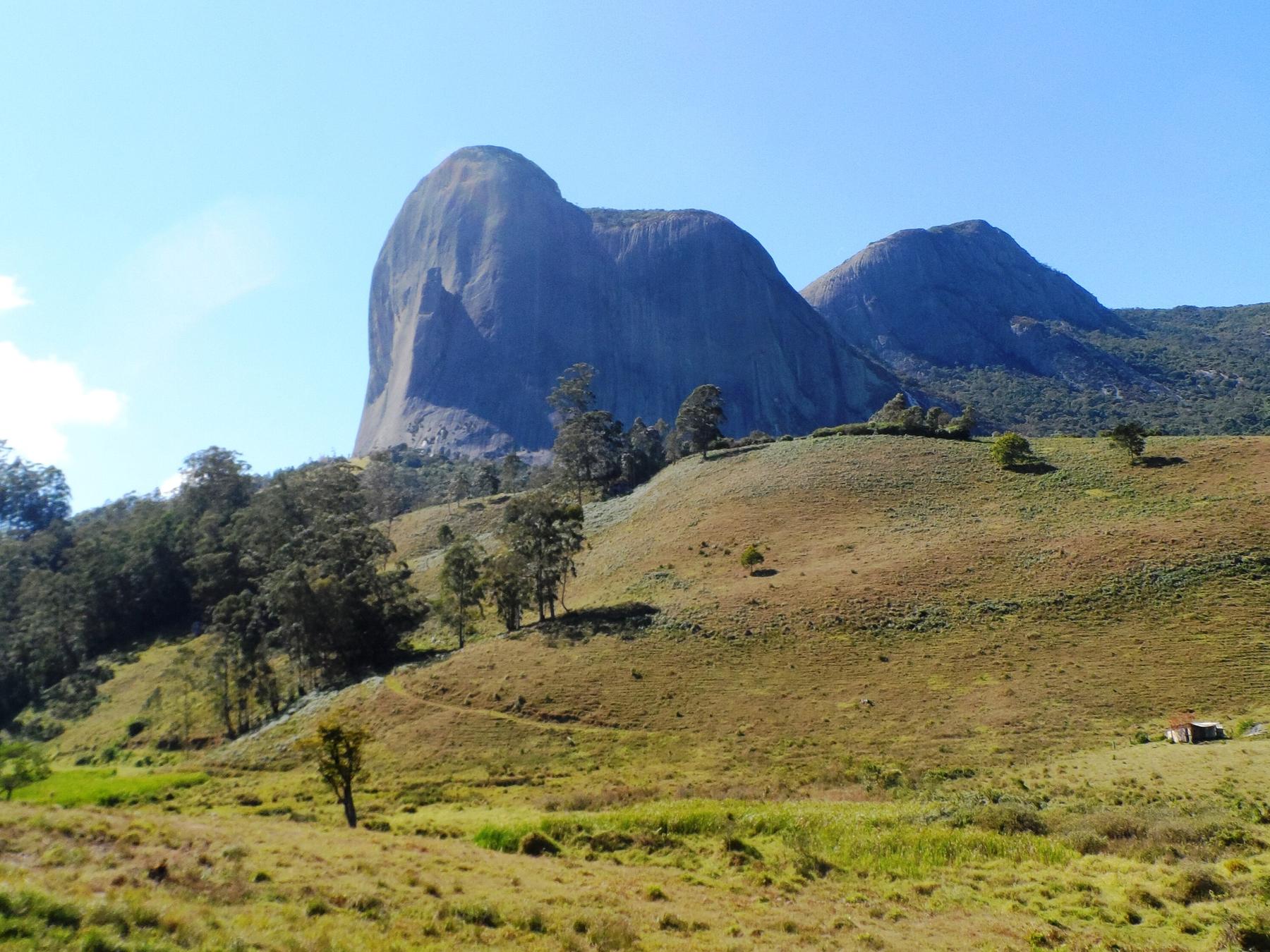 Pedra Azul e a Pedra do Lagarto vistas de um campo de pastagem verde em Domingos Martins, com os paredões de granito cinza contra o céu azul.