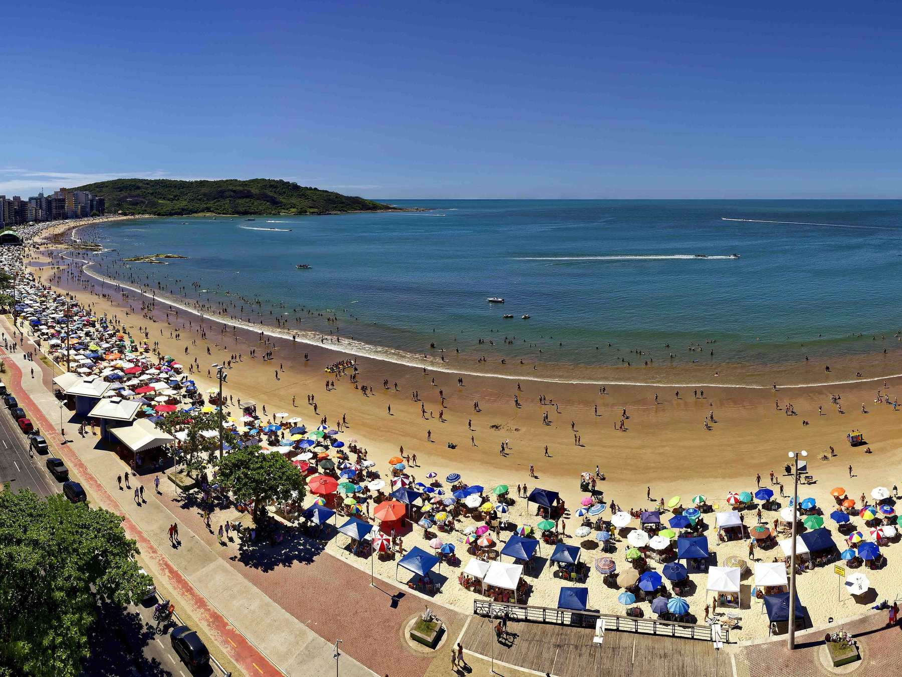 Praia do Morro vista do alto em Guarapari, com a curva da areia tomada por guarda-sóis coloridos, o calçadão movimentado e o mar azul.