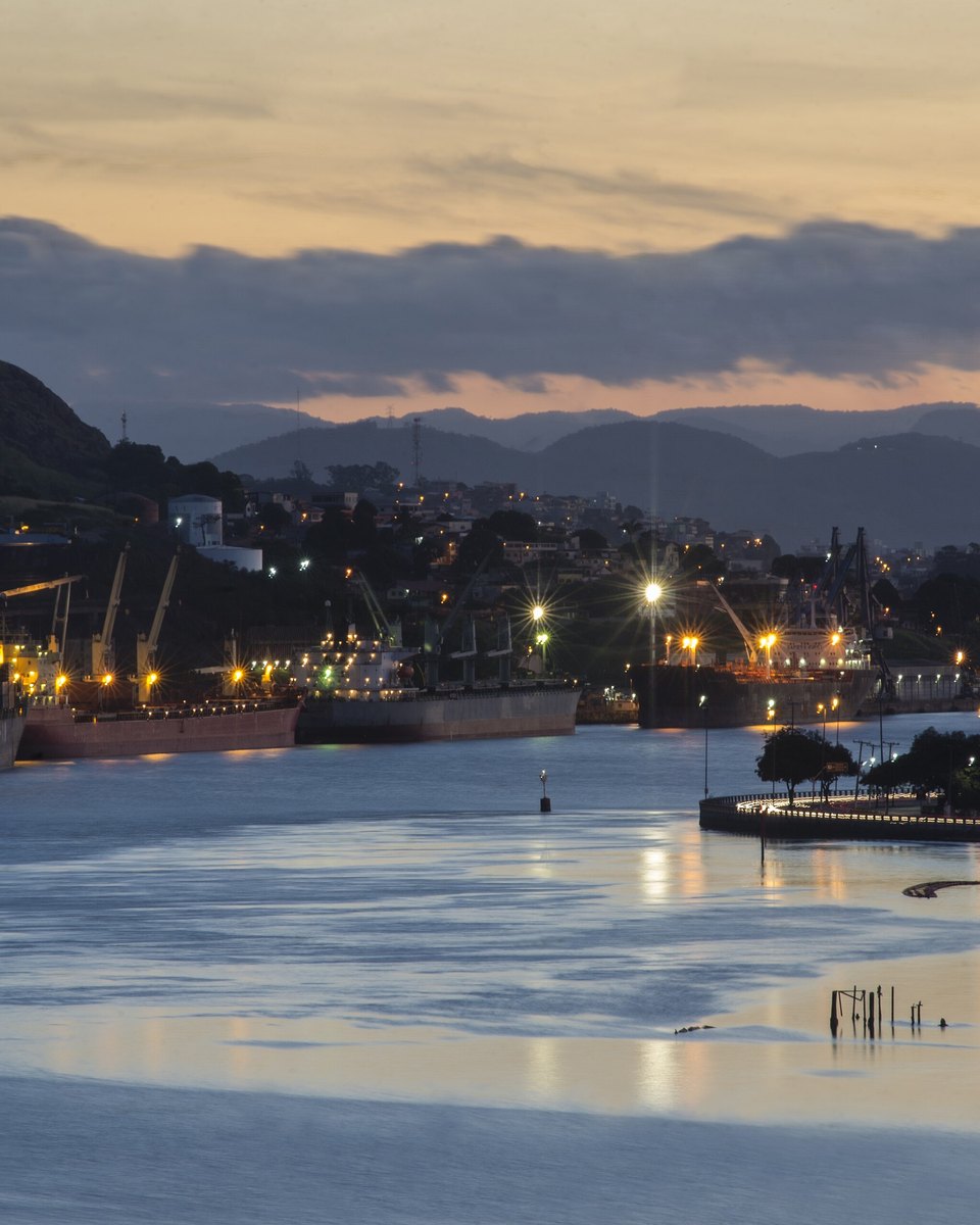 Navios de carga e guindastes no Porto de Vitória ao entardecer, com as luzes da cidade refletidas na água.