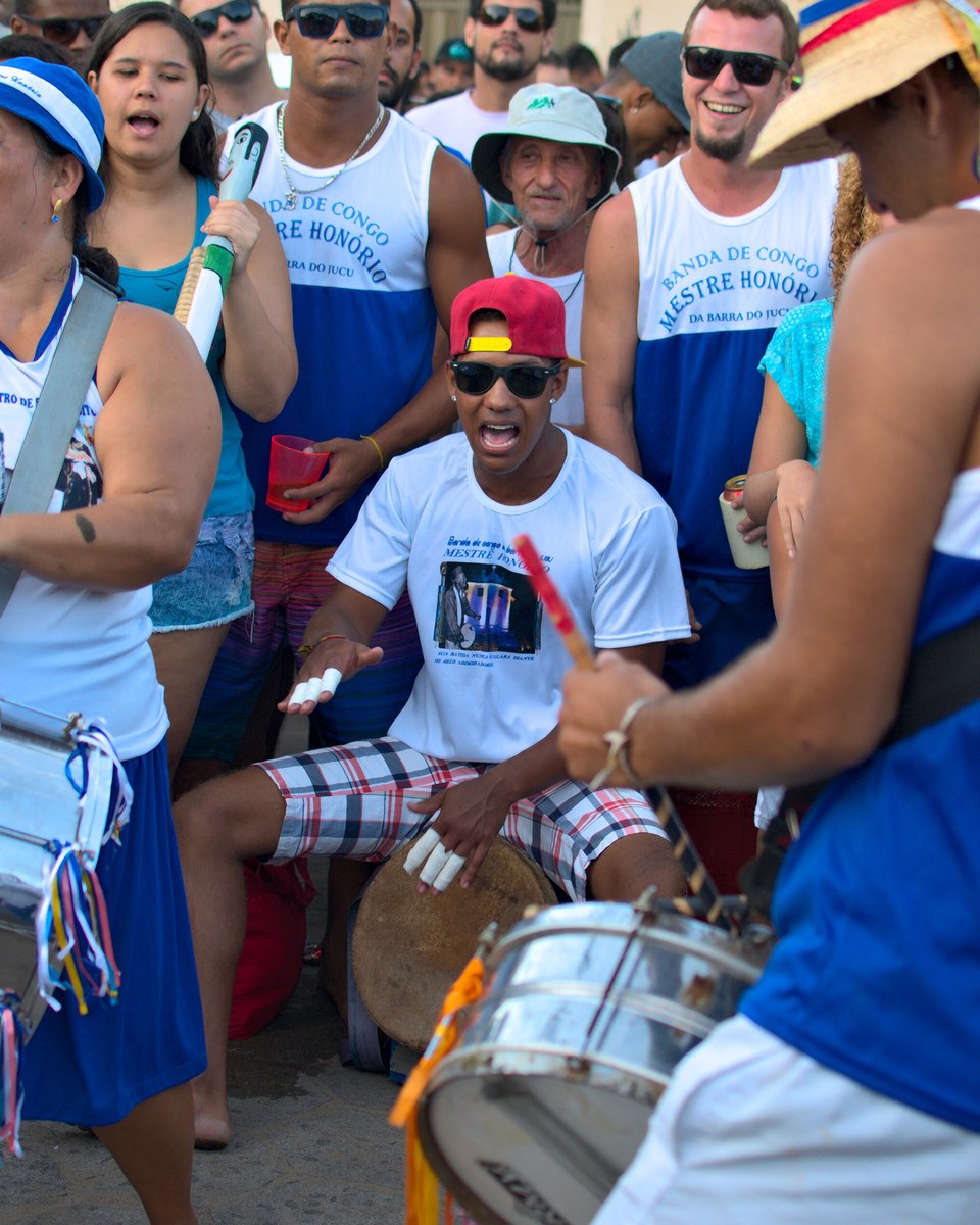 Integrantes de banda de congo tocando tambores e cantando em apresentação de rua, de uniforme azul e branco.