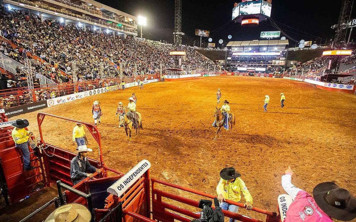 Arena da Festa do Peão de Barretos à noite, com a arquibancada lotada e peões a cavalo na areia.