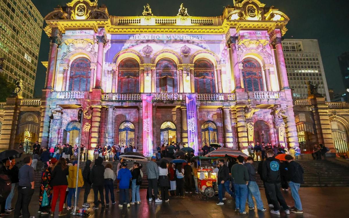 Theatro Municipal de São Paulo iluminado com projeções e banners da Virada Cultural, com o público na escadaria à noite.