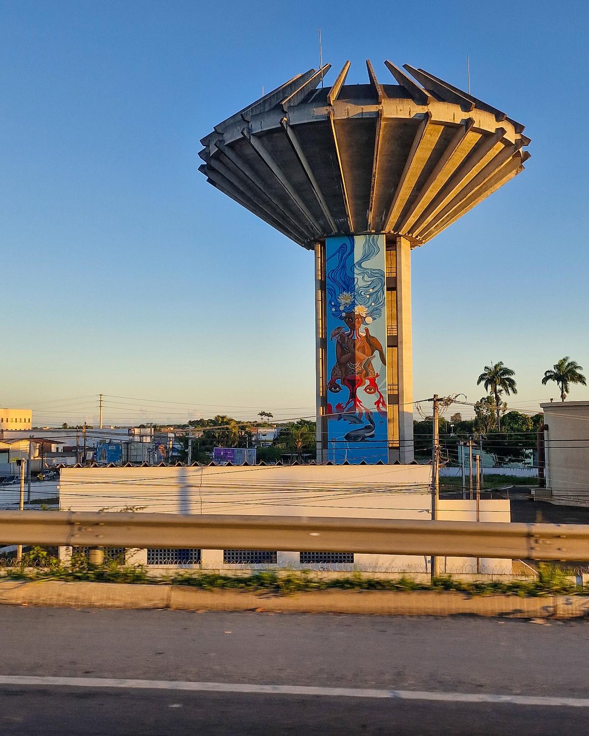 Caixa d'água do Tomba, torre de concreto com o reservatório em forma de taça no alto e uma pintura colorida no pilar, na luz do fim de tarde sob céu azul.