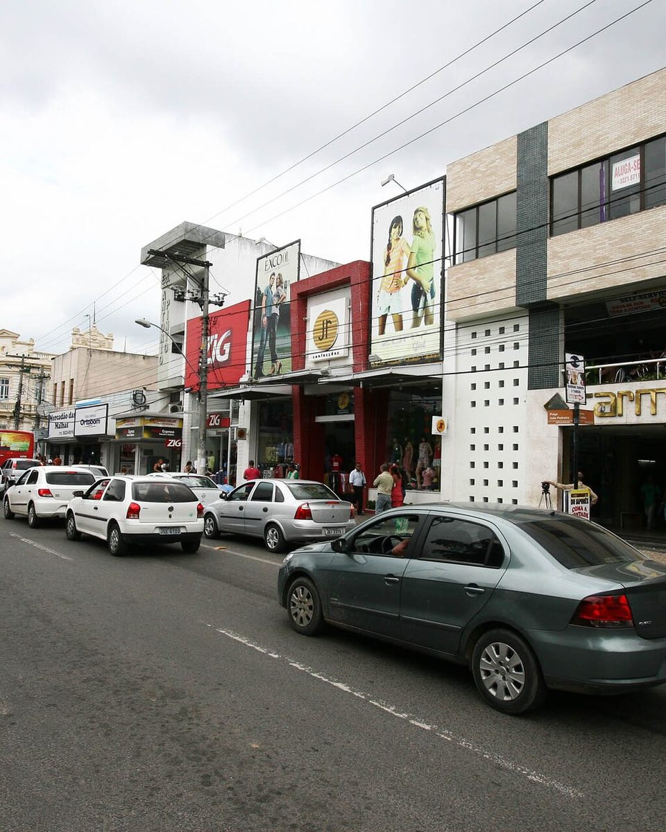 Rua comercial do Centro de Feira de Santana com lojas de fachada colorida, placas, carros e motos passando e pedestres na calçada, num dia nublado.