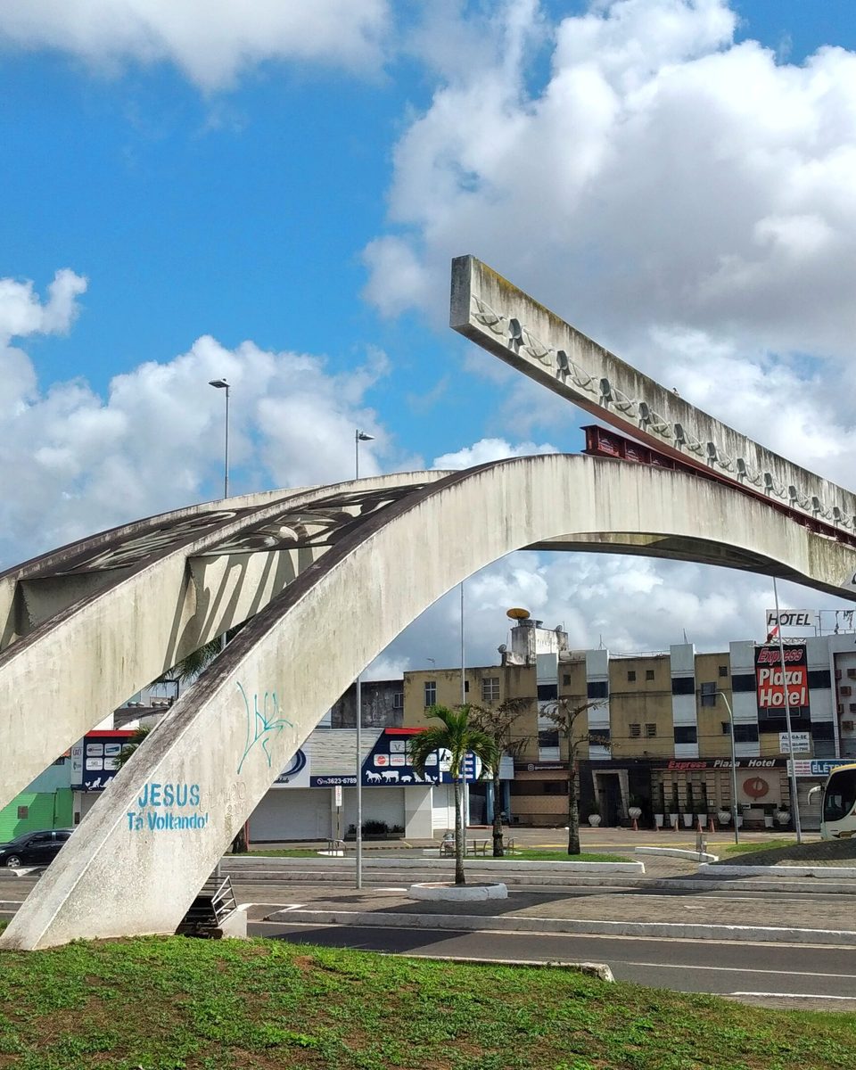 Monumento ao Caminhoneiro, em Feira de Santana, com arcos de concreto claro sobre um canteiro gramado, palmeiras em volta, lojas ao fundo e céu azul com nuvens.