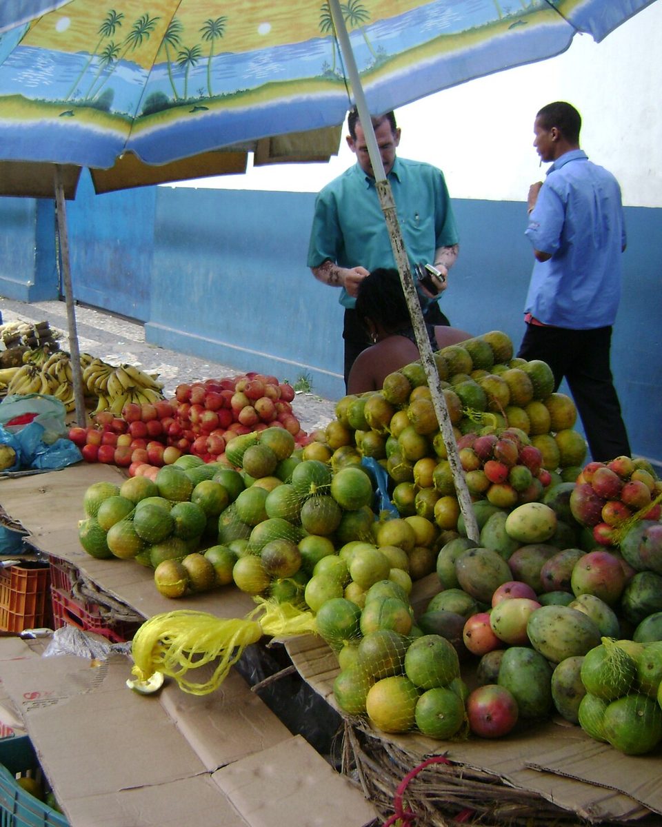 Banca de feira em Feira de Santana com pilhas de mangas e outras frutas sob guarda-sóis coloridos, uma feirante arrumando a mercadoria e dois homens conversando ao lado.