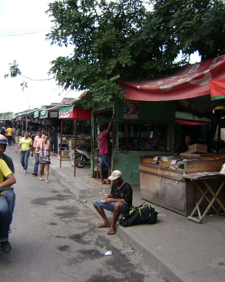 Rua de feira livre em Feira de Santana com barracas de lona vermelha na calçada, um motociclista de capacete passando com um garupa, carros estacionados e pessoas caminhando entre as bancas.