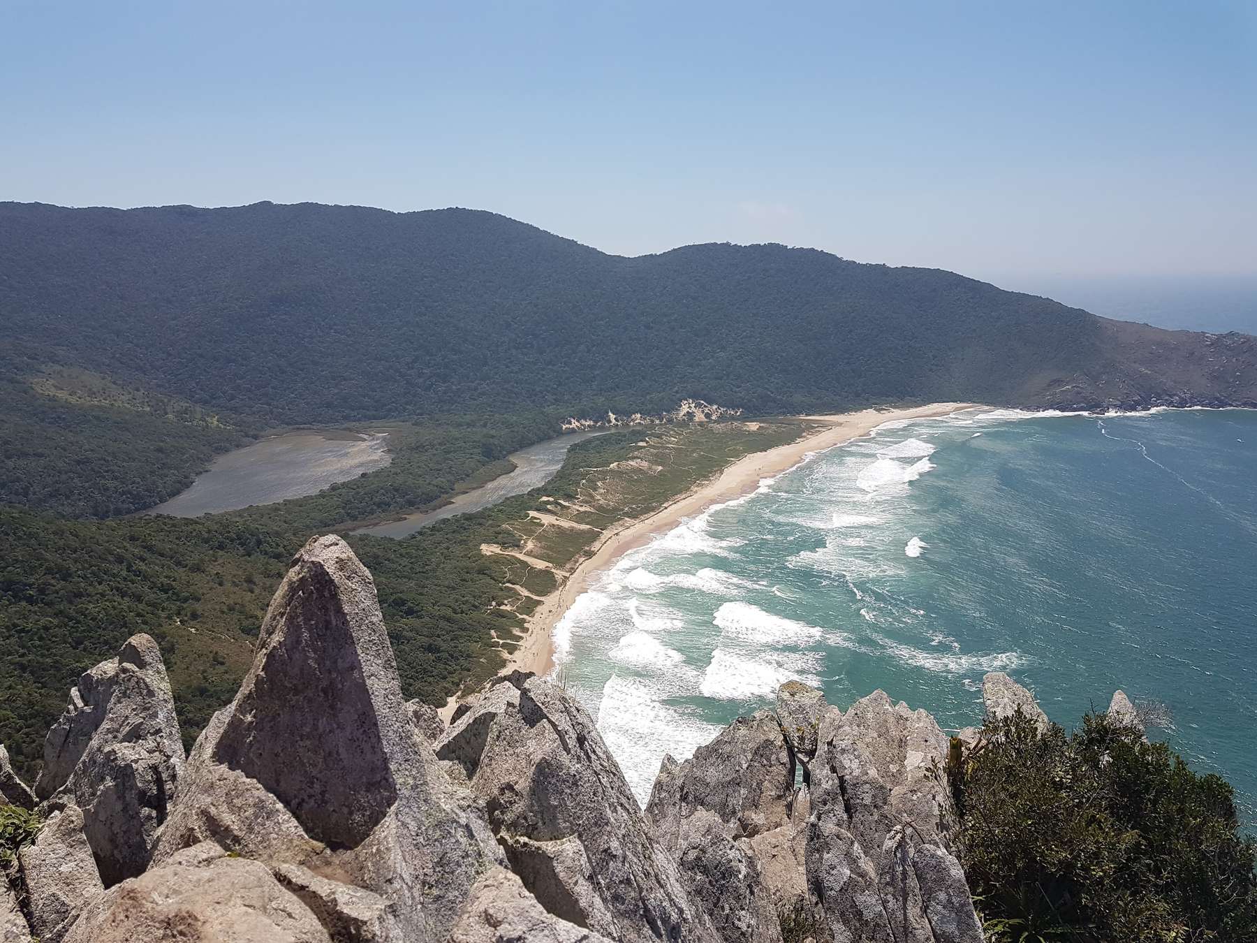 Praia da Lagoinha do Leste vista do alto do morro, entre pedras pontiagudas em primeiro plano, com a lagoa atrás da areia, a mata descendo até o mar e as ondas brancas quebrando.