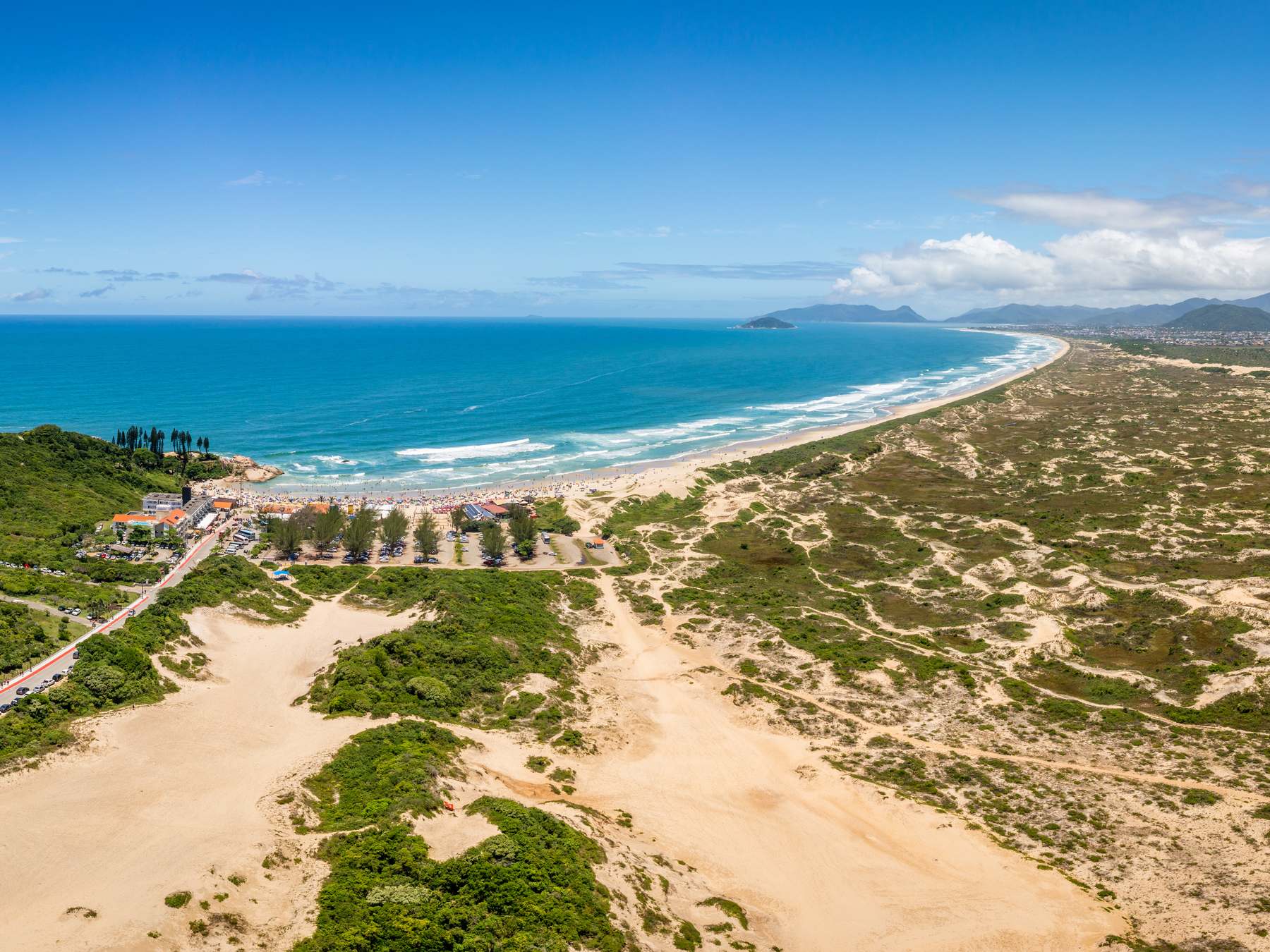 Dunas da Joaquina vistas do alto, com a areia clara entre a vegetação de restinga, a estrada que chega à praia e o mar azul com a Praia da Joaquina e o Campeche ao fundo.