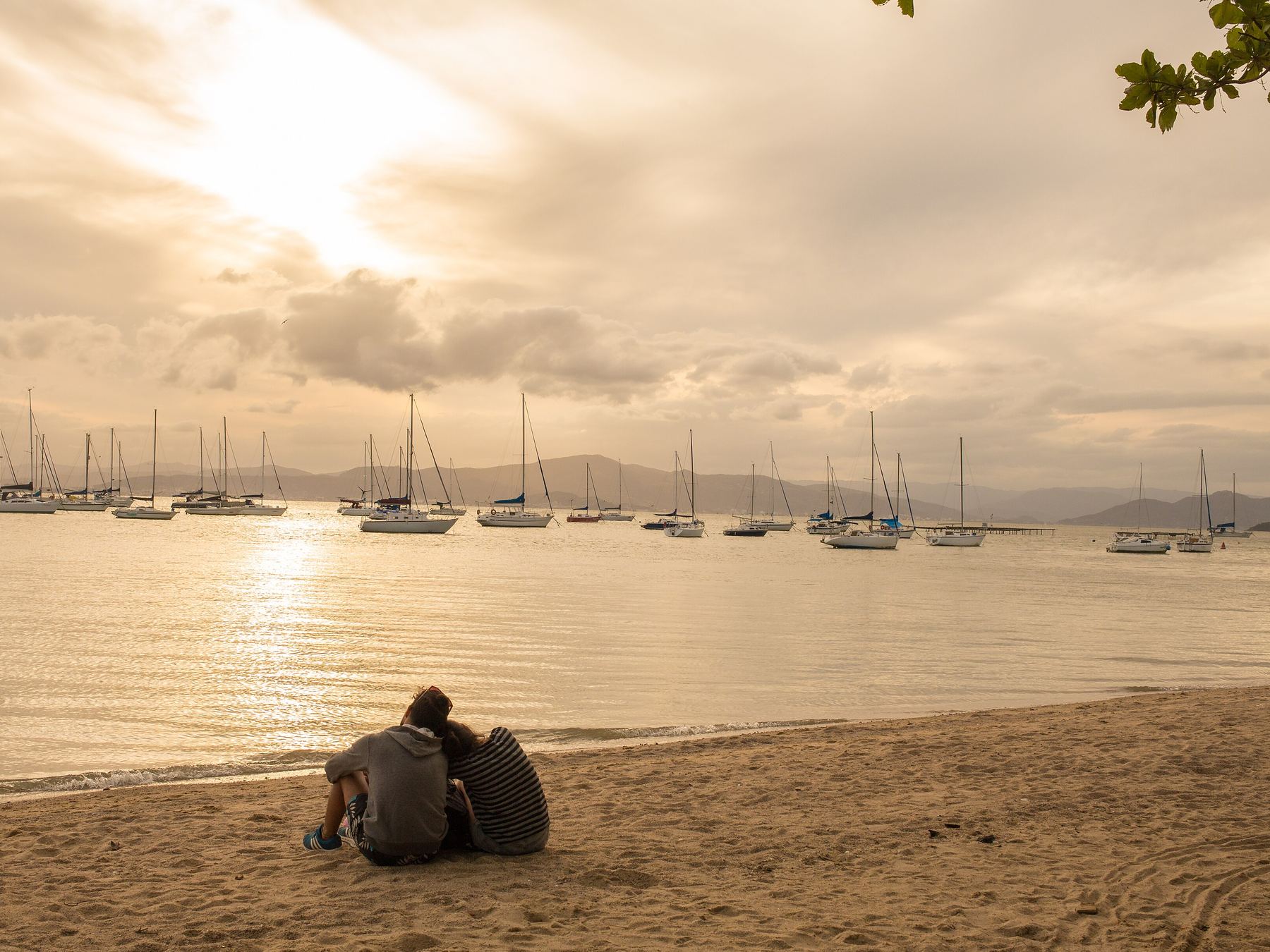 Casal sentado na areia da praia de Santo Antônio de Lisboa ao pôr do sol, olhando os veleiros ancorados na baía dourada sob um céu de nuvens alaranjadas.