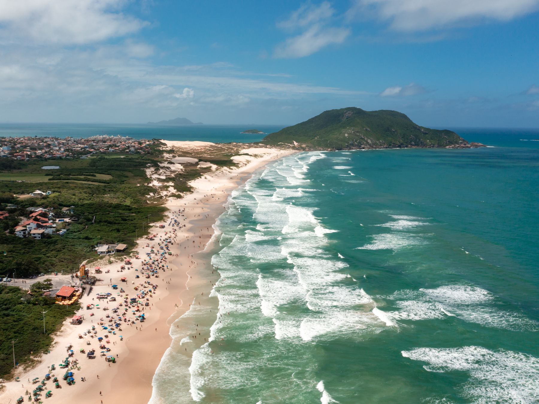 Praia do Santinho vista do alto num dia de sol, com a faixa de areia cheia de banhistas, as ondas brancas em sequência no mar verde-azulado e o morro do costão ao fundo.