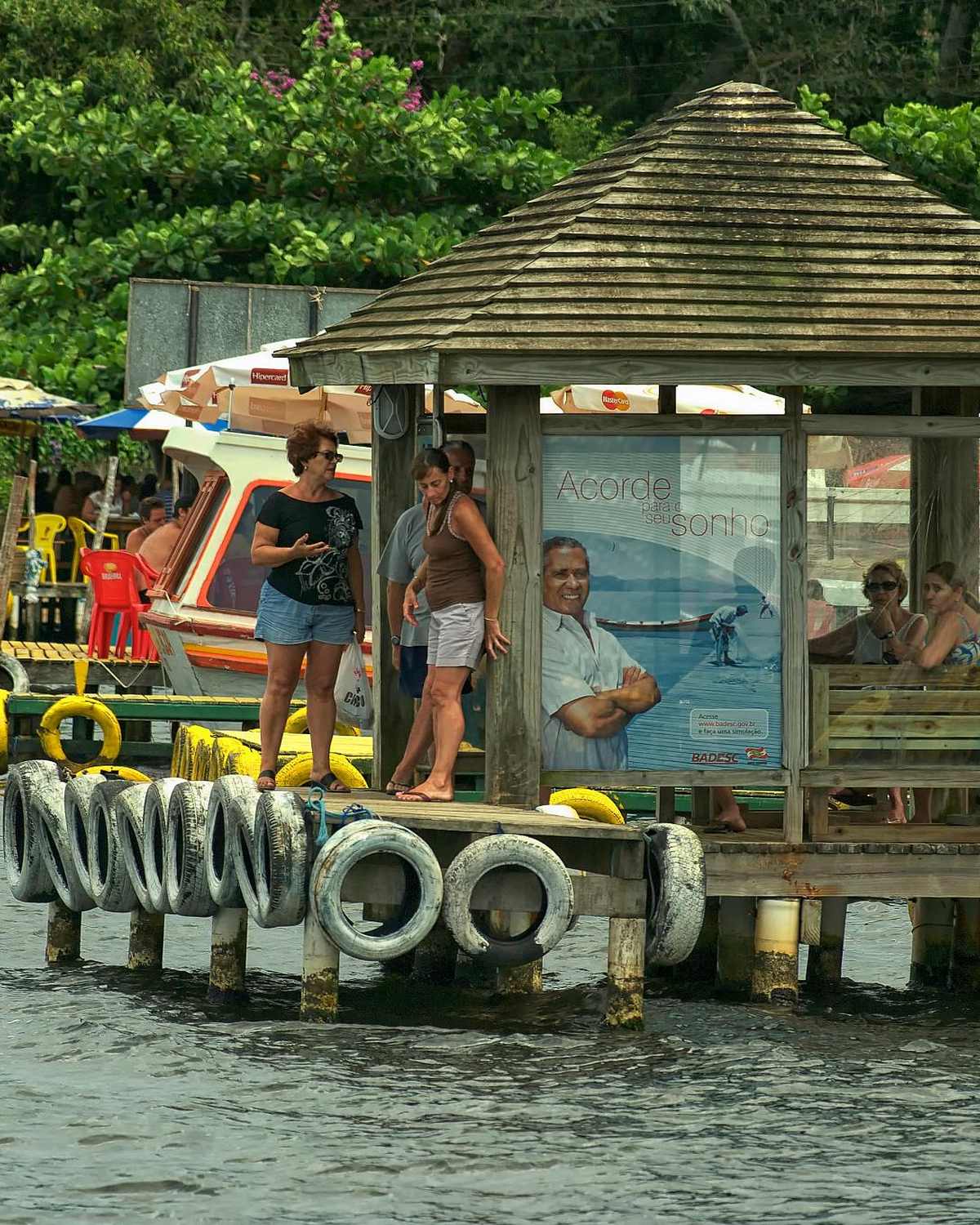 Moradores e visitantes num trapiche de madeira da Costa da Lagoa, em Florianópolis, à espera do barco, com pneus pendurados na borda, barcos coloridos e a mata logo atrás.