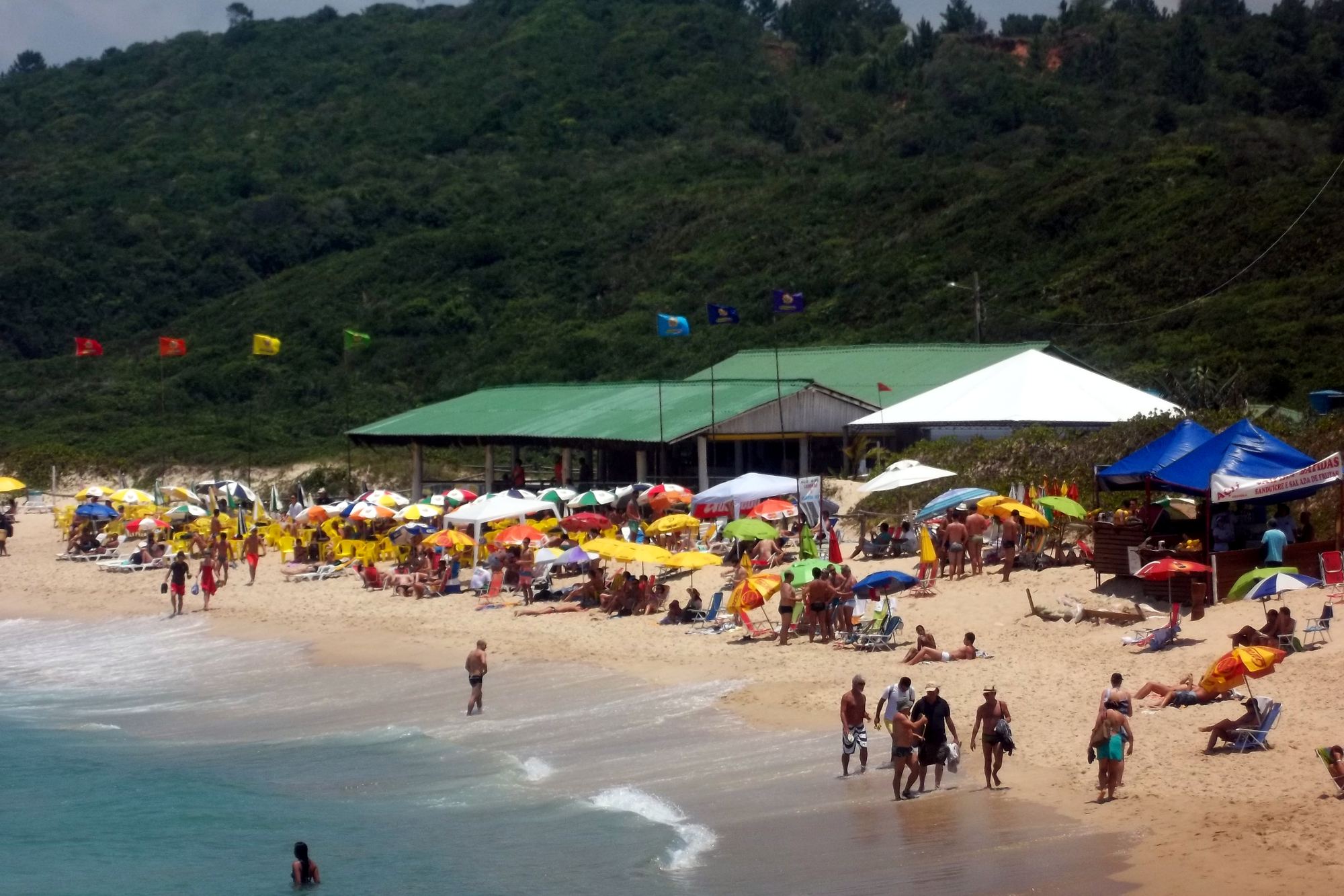 Praia Mole cheia num dia de sol de verão, em Florianópolis, com guarda-sóis coloridos na areia, gente caminhando e entrando no mar e o morro coberto de mata atrás do bar de teto verde.