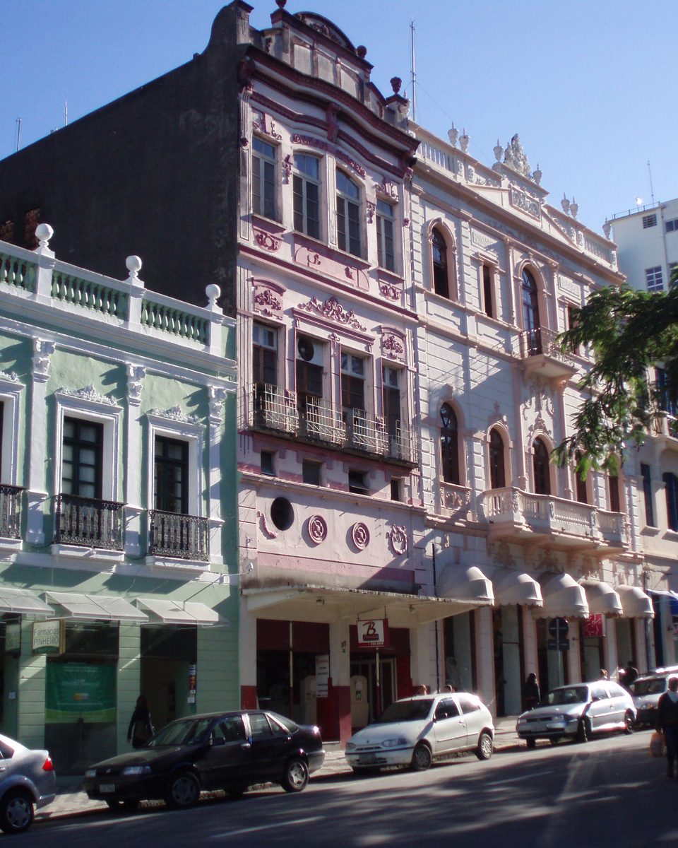 Fileira de casarões históricos coloridos no Centro de Florianópolis, com sacadas de ferro, fachadas verde-clara, rosa e branca, carros estacionados e pedestres na calçada sob céu azul.