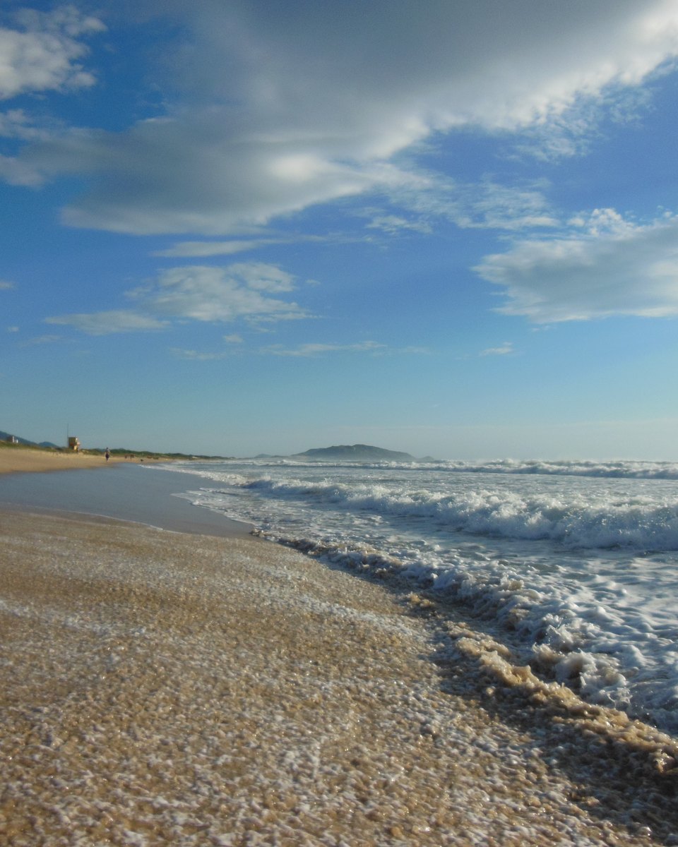 Beira da água na Praia do Campeche, em Florianópolis, de manhã cedo, com a espuma das ondas chegando à areia, ilhas no horizonte e nuvens brancas no céu azul.