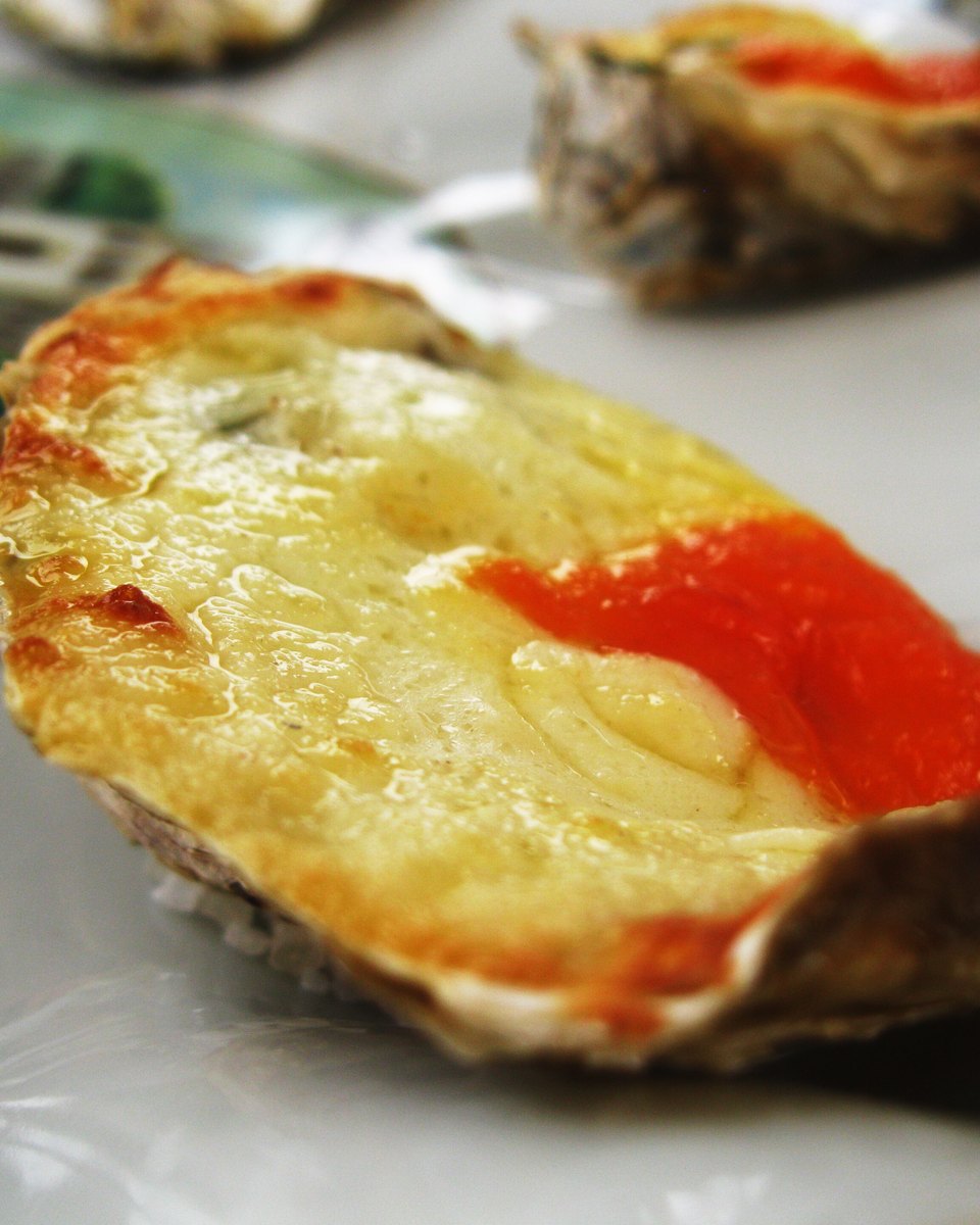 Ostra gratinada com queijo dourado e molho vermelho servida na concha, num prato branco de restaurante em Florianópolis, com outras ostras ao fundo.