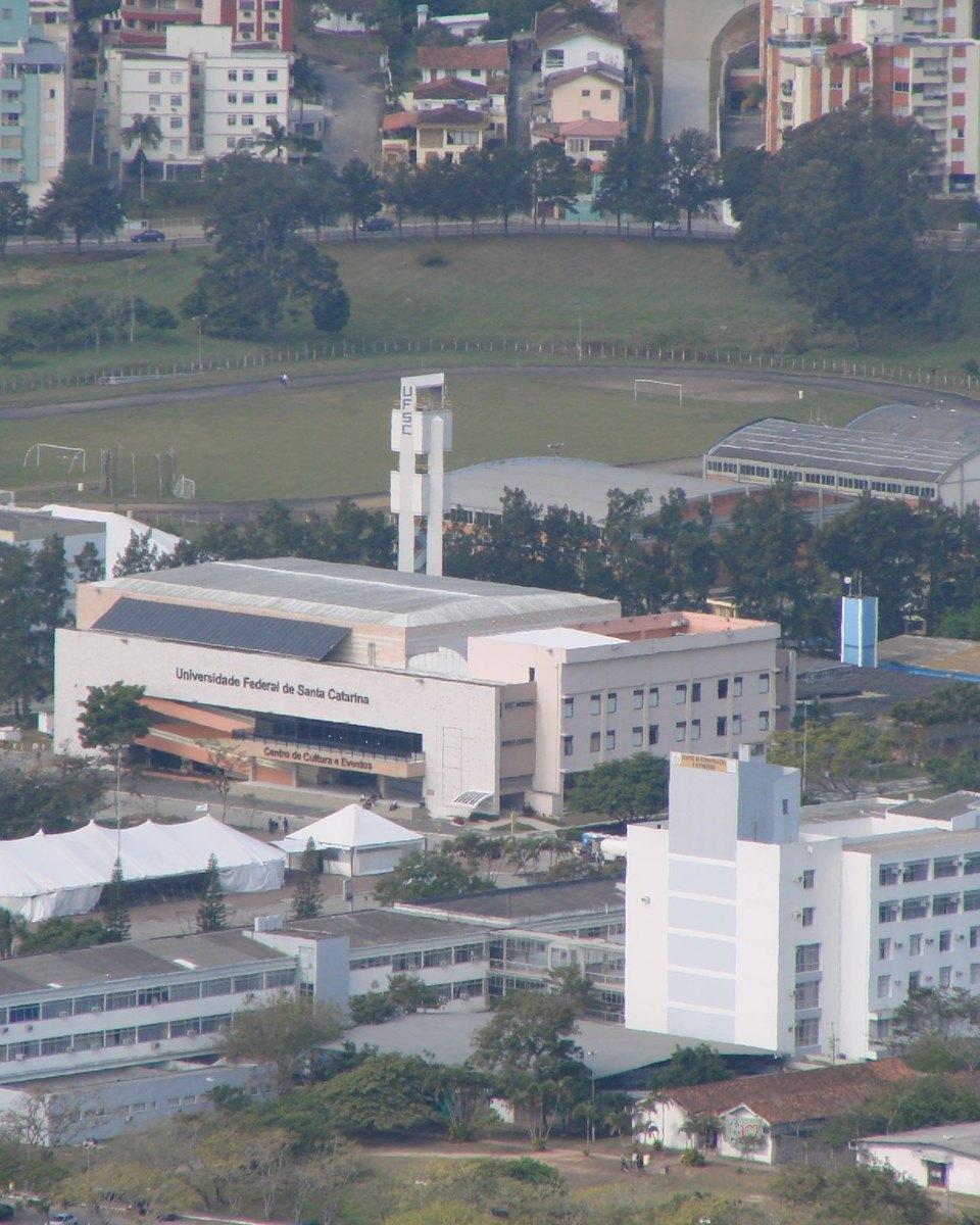 Vista do alto de parte do campus da UFSC, na Trindade, em Florianópolis, com o Centro de Cultura e Eventos no meio, prédios brancos da universidade entre árvores e os edifícios residenciais do bairro ao fundo.