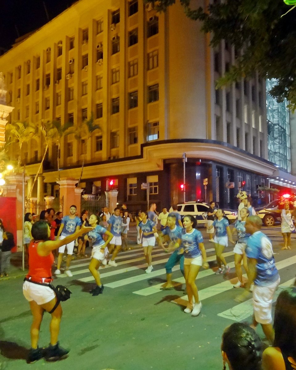 Bloco de Carnaval passando à noite por uma rua do Centro de Florianópolis, com passistas de camiseta azul dançando na faixa de pedestres, público nas calçadas e um prédio iluminado ao fundo.