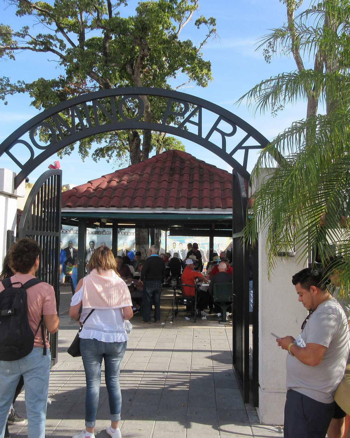 Pessoas entrando pelo portão em arco do Domino Park, em Little Havana, onde moradores jogam dominó sob um quiosque de telhado vermelho.