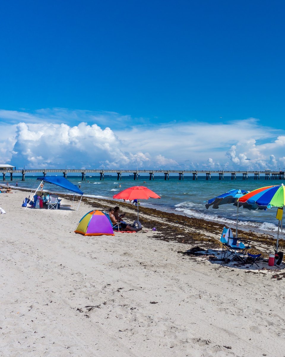 Guarda-sóis coloridos e barracas na areia de Dania Beach, no condado de Broward, com banhistas no mar e o píer ao fundo.