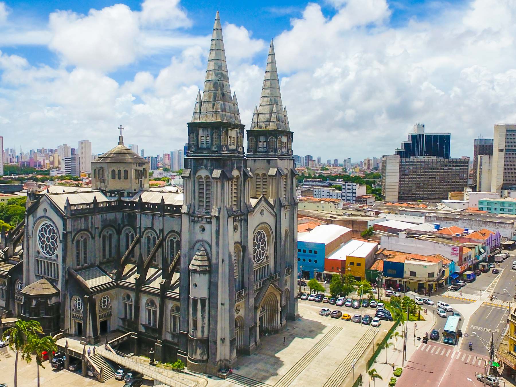 Catedral Metropolitana de Fortaleza vista do alto, com as duas torres góticas de pedra cinza, o centro da cidade em volta e o céu azul com nuvens brancas.