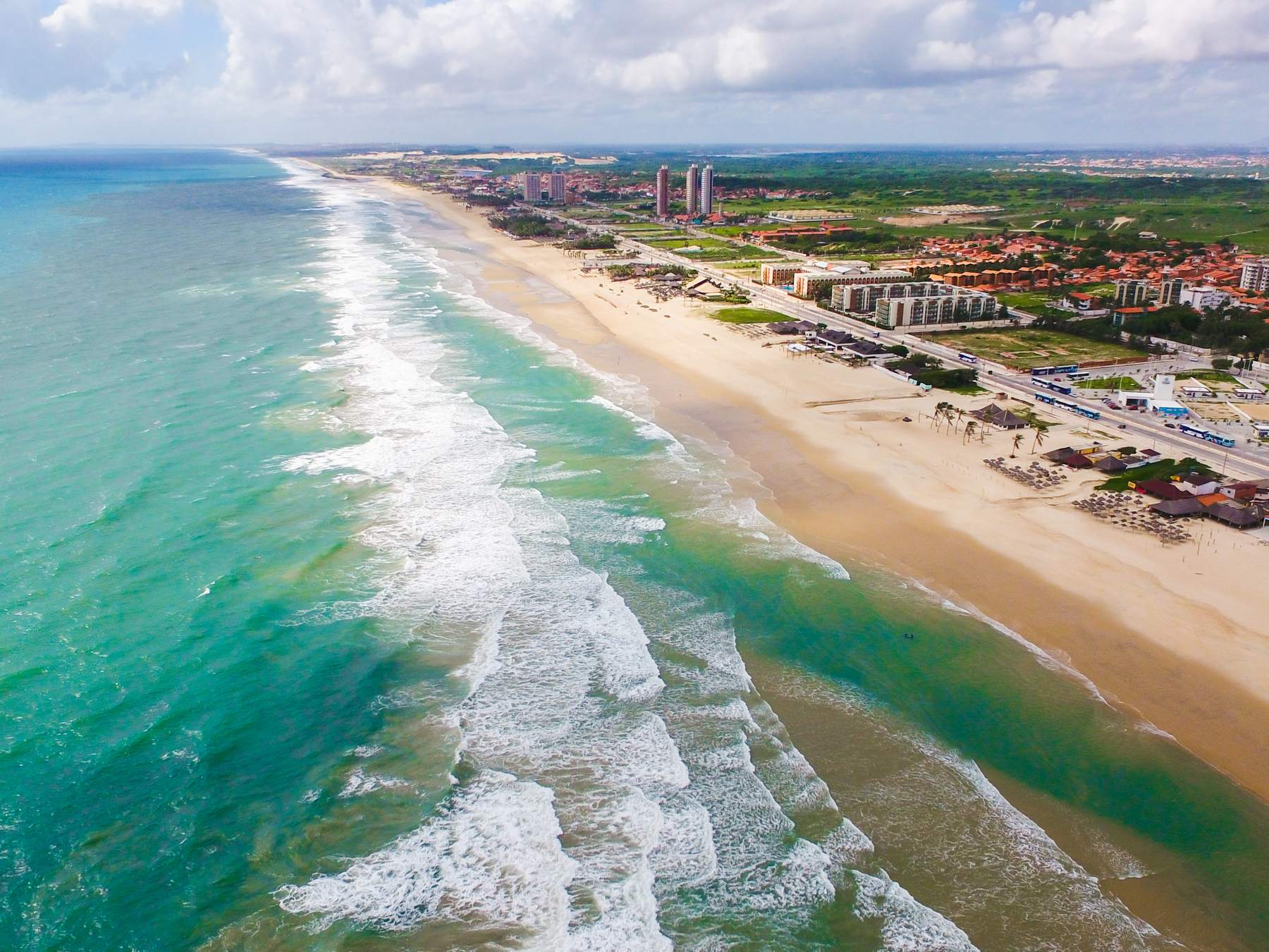 Vista aérea da Praia do Futuro, em Fortaleza, com o mar verde-turquesa, as ondas brancas quebrando em linhas e a faixa longa de areia com as barracas até o horizonte.