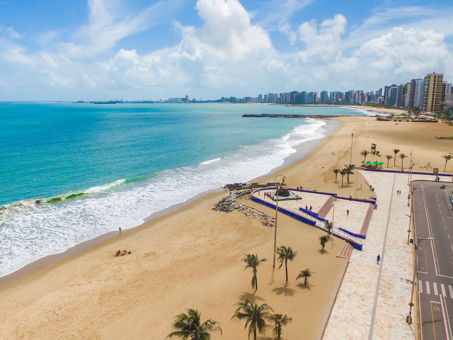 Vista aérea da Praia de Iracema, em Fortaleza, com o mar azul e as ondas brancas, a areia dourada, os coqueiros do calçadão e a fileira de prédios da orla ao fundo sob nuvens brancas.