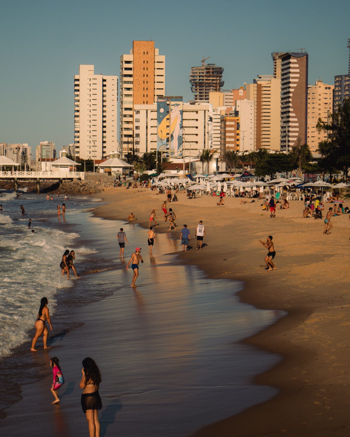 Pessoas caminhando e entrando no mar na beira da Praia de Iracema, em Fortaleza, na luz dourada do fim da tarde, com a areia molhada refletindo o céu e os prédios da orla ao fundo.
