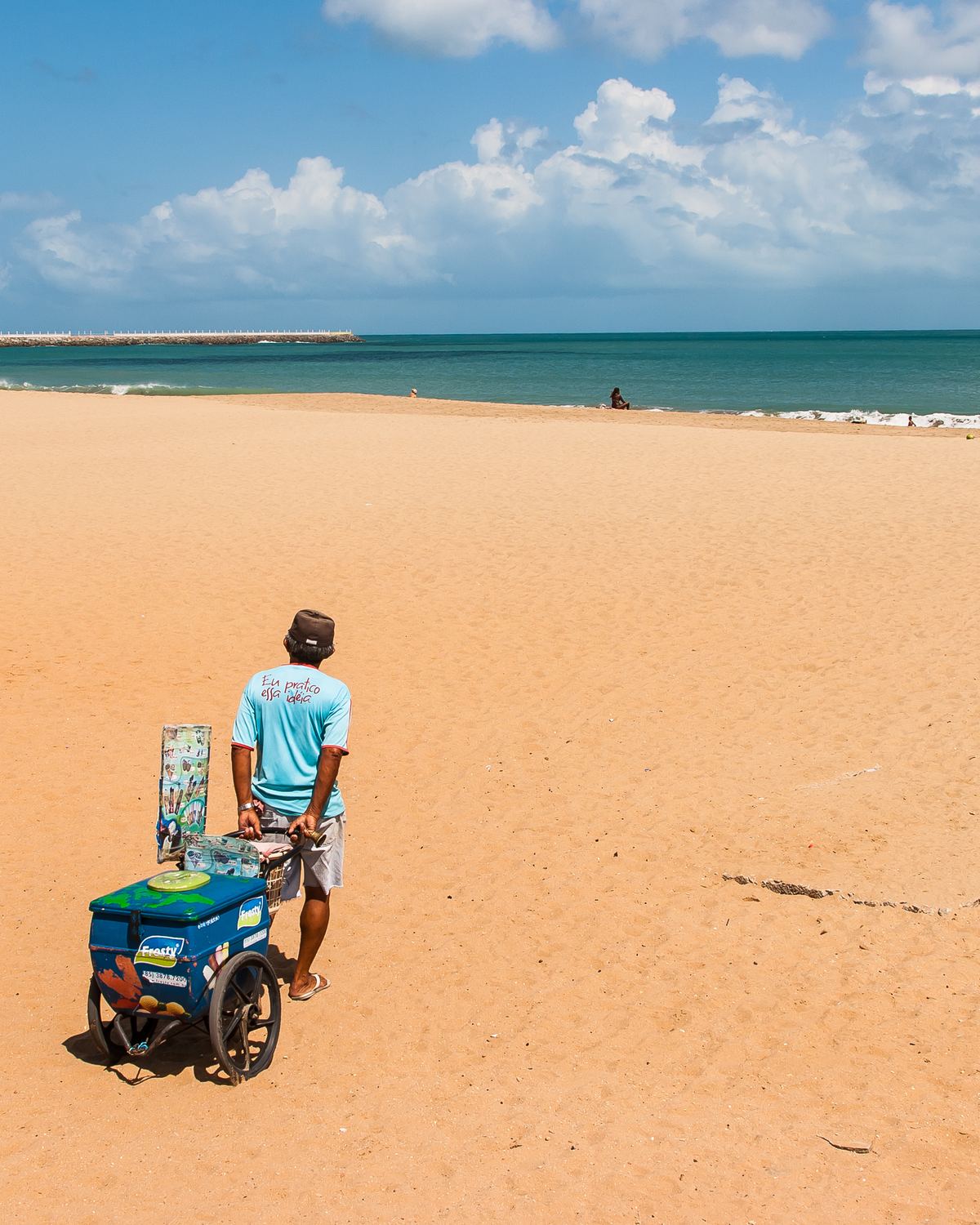 Vendedor de sorvete empurrando o carrinho pela areia clara e fofa da orla de Fortaleza, com o mar verde, o quebra-mar e o céu azul com nuvens ao fundo.