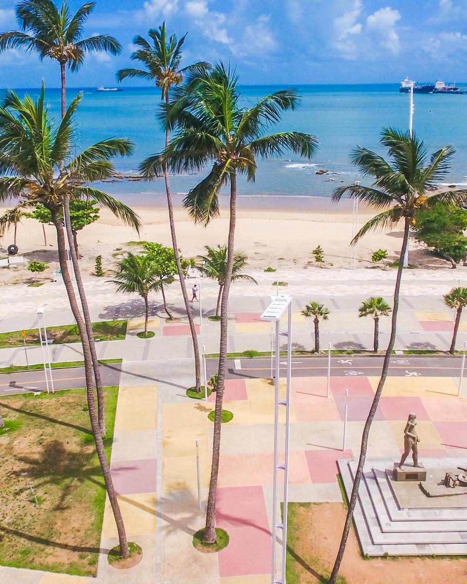 Estátua de Iracema Guardiã vista do alto na Praia de Iracema, sobre um piso colorido, entre coqueiros, com a areia, as pedras e o mar azul ao fundo.