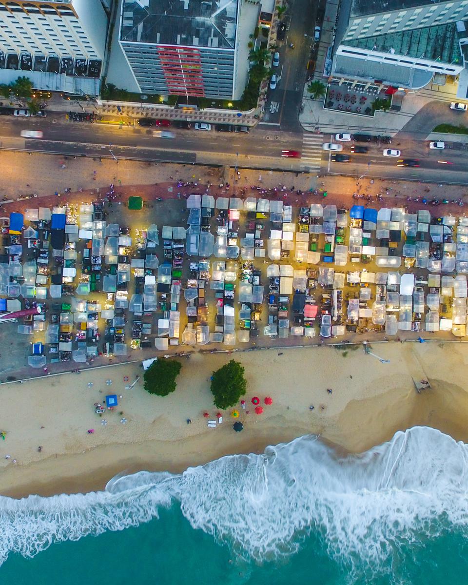 Vista aérea da feirinha da Beira-Mar no fim da tarde, com centenas de barracas de lona colorida enfileiradas entre a avenida e a praia, as ondas quebrando na areia e os prédios iluminados.